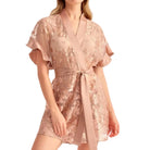 A woman modeling a short, sheer floral lace robe dress in a dusty rose pink color, featuring flutter sleeves and a matching tie belt, set against a plain light gray studio background.
