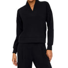A model wearing a black long-sleeve top with a unique folded collar and matching black trousers, photographed against a solid off-white studio background.
