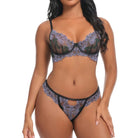 A woman modeling a two-piece lace lingerie set with a floral pattern in muted purple and green tones, accented by black trim, against a plain white background.