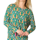 A woman wearing a long-sleeve pajama top with a green base and a colorful paisley and floral print, paired with matching pants, against a plain light background.