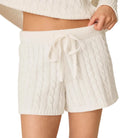 A person wearing cream cable-knit shorts with a matching drawstring waistband, paired with a cropped cream cable-knit sweater, shown against a plain light background.