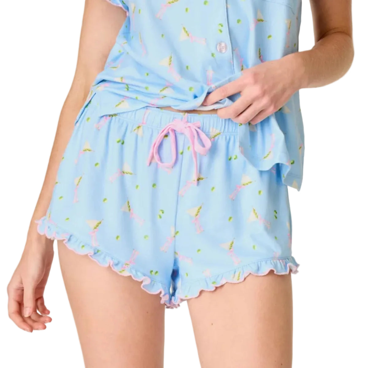 A model wearing a light blue pajama short with a pastel blue martini print, featuring a short-sleeve top and matching ruffled shorts with a pink drawstring, photographed against a solid off-white background.