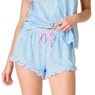A model wearing a light blue pajama short with a pastel blue martini print, featuring a short-sleeve top and matching ruffled shorts with a pink drawstring, photographed against a solid off-white background.