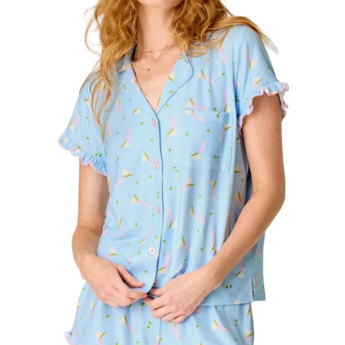 A woman modeling a light blue pajama set with a small floral print, featuring a short-sleeve button-front top and matching shorts, against a plain off-white studio background.