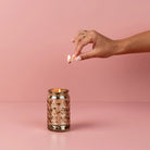 Hand lighting a candle in a decorative jar against a pink background