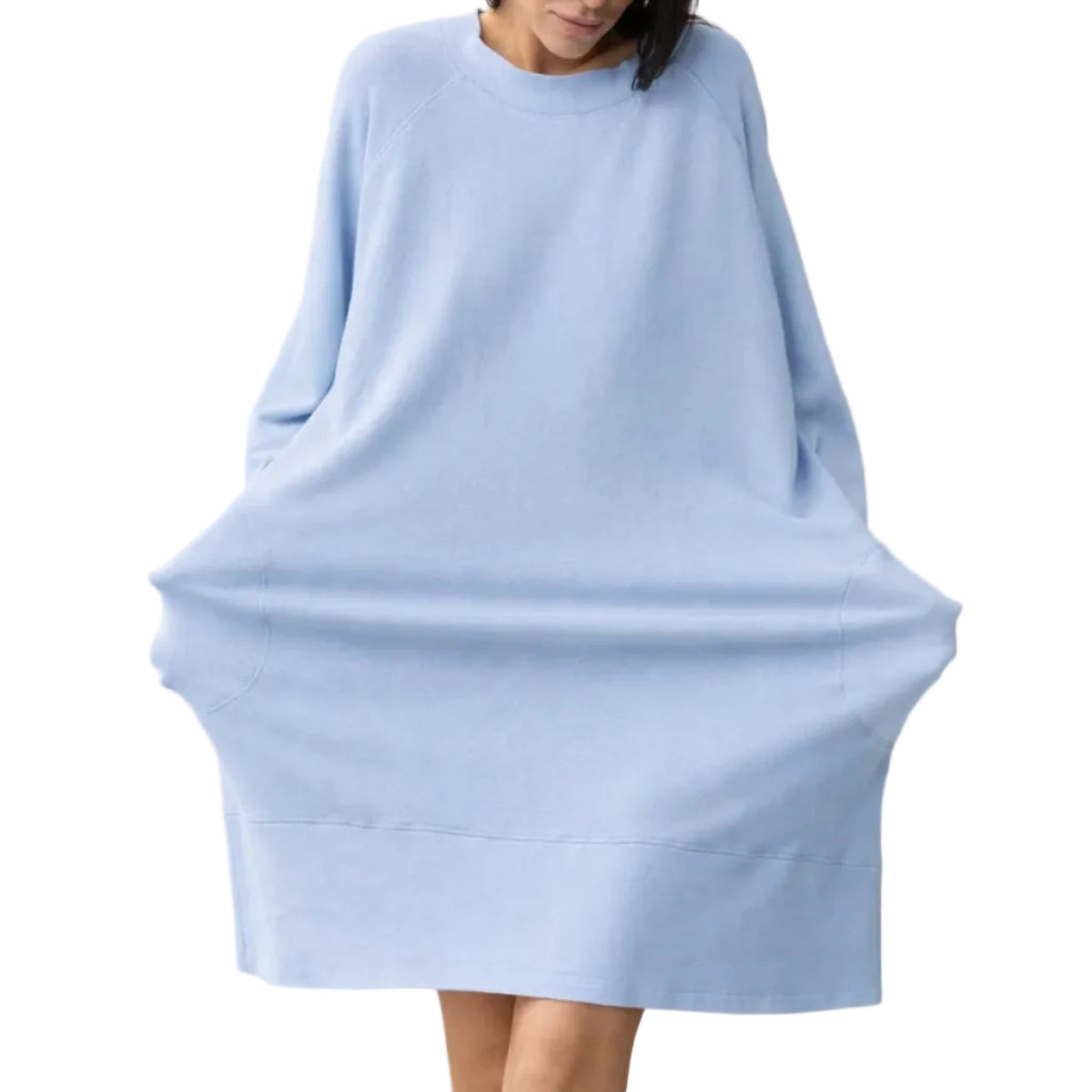A woman wearing a light blue oversized long-sleeve knit top or poncho-style sweater, standing against a plain beige studio background.